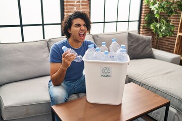 Canvas Print - Hispanic man with curly hair holding recycling bin with plastic bottles at home angry and mad screaming frustrated and furious, shouting with anger. rage and aggressive concept.