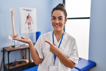 Poster - Young hispanic woman working at rehabilitation clinic amazed and smiling to the camera while presenting with hand and pointing with finger.