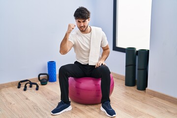 Canvas Print - Hispanic man with beard sitting on pilate balls at yoga room angry and mad raising fist frustrated and furious while shouting with anger. rage and aggressive concept.