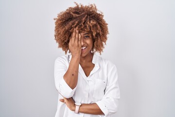Young hispanic woman with curly hair standing over white background covering one eye with hand, confident smile on face and surprise emotion.