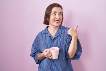 Poster - Middle age hispanic woman drinking a cup coffee smiling with happy face looking and pointing to the side with thumb up.