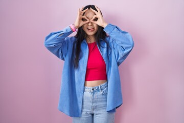 Sticker - Young asian woman standing over pink background doing ok gesture like binoculars sticking tongue out, eyes looking through fingers. crazy expression.