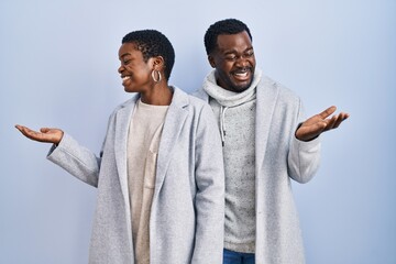 Poster - Young african american couple standing over blue background together smiling showing both hands open palms, presenting and advertising comparison and balance