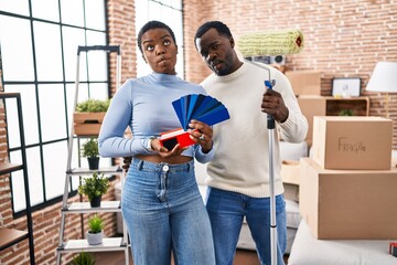 Canvas Print - Young african american couple moving to a new home choosing walls color making fish face with mouth and squinting eyes, crazy and comical.