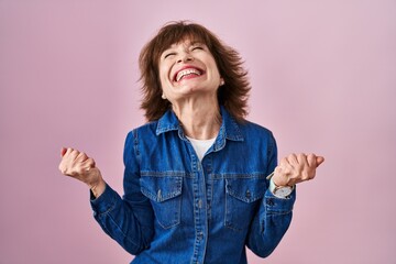 Wall Mural - Middle age woman standing over pink background celebrating surprised and amazed for success with arms raised and eyes closed. winner concept.