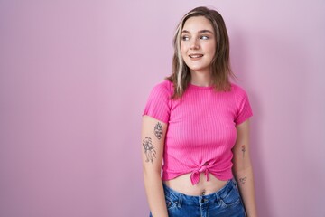 Wall Mural - Blonde caucasian woman standing over pink background looking away to side with smile on face, natural expression. laughing confident.