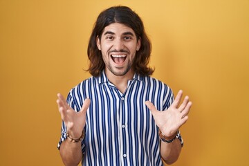 Wall Mural - Hispanic man with long hair standing over yellow background crazy and mad shouting and yelling with aggressive expression and arms raised. frustration concept.