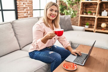 Wall Mural - Young hispanic woman using laptop and drinking coffee sitting on sofa at home