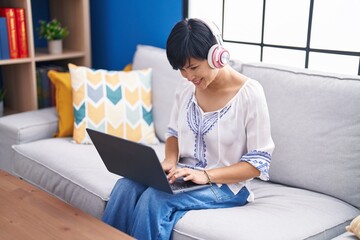 Wall Mural - Middle age chinese woman using laptop and headphones sitting on sofa at home