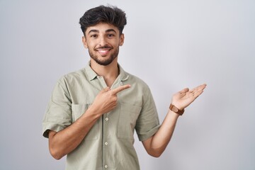 Poster - Arab man with beard standing over white background amazed and smiling to the camera while presenting with hand and pointing with finger.