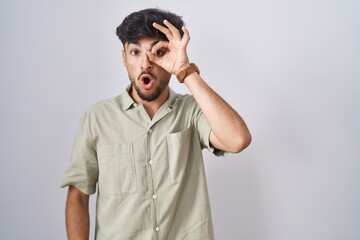 Canvas Print - Arab man with beard standing over white background doing ok gesture shocked with surprised face, eye looking through fingers. unbelieving expression.