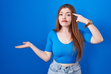 Poster - Redhead woman standing over blue background confused and annoyed with open palm showing copy space and pointing finger to forehead. think about it.