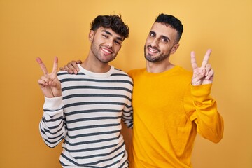 Wall Mural - Young hispanic gay couple standing over yellow background smiling looking to the camera showing fingers doing victory sign. number two.