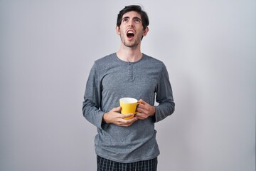 Poster - Young hispanic man wearing pajama drinking a cup of coffee angry and mad screaming frustrated and furious, shouting with anger. rage and aggressive concept.