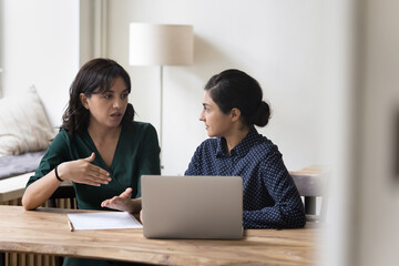 Canvas Print - Two multi-ethnic businesswomen colleagues sit at desk, discuss joint project sit in office at workplace desk with laptop, share vision, ideas, planning cooperation. Teamwork, apprenticeship, mentoring