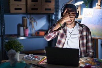 Poster - Young hispanic man sitting at art studio with laptop late at night smiling and laughing with hand on face covering eyes for surprise. blind concept.