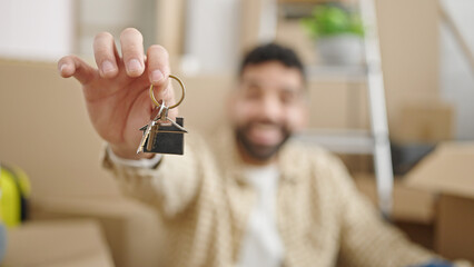 Young hispanic man holding keys sitting on floor at new home