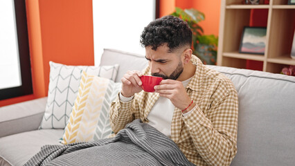 Sticker - Young hispanic man drinking coffee sitting on sofa at h