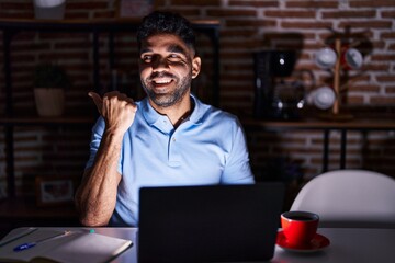 Canvas Print - Hispanic man with beard using laptop at night smiling with happy face looking and pointing to the side with thumb up.