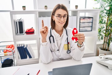 Sticker - Young caucasian doctor woman holding support red ribbon surprised with an idea or question pointing finger with happy face, number one