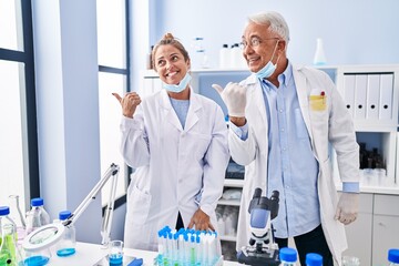 Poster - Middle age hispanic people working at scientist laboratory pointing thumb up to the side smiling happy with open mouth