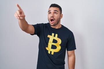 Poster - Young hispanic man wearing bitcoin t shirt pointing with finger surprised ahead, open mouth amazed expression, something on the front