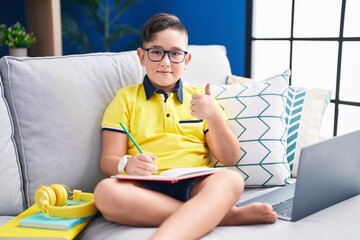 Poster - Young hispanic kid doing homework sitting on the sofa doing happy thumbs up gesture with hand. approving expression looking at the camera showing success.