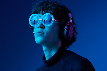 Wall Mural - Teenage male with headphones listening to music and dancing and singing with glasses, hipster lifestyle, blue background, neon light, style and trends, mixed light, copy space