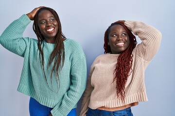 Poster - Two african woman standing over blue background smiling confident touching hair with hand up gesture, posing attractive and fashionable