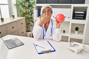 Poster - Middle age woman with grey hair wearing doctor uniform holding balloon covering mouth with hand, shocked and afraid for mistake. surprised expression