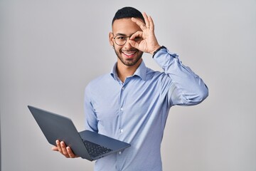 Poster - Young hispanic man working using computer laptop doing ok gesture with hand smiling, eye looking through fingers with happy face.