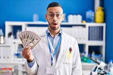 Poster - Young hispanic man working at scientist laboratory holding money scared and amazed with open mouth for surprise, disbelief face