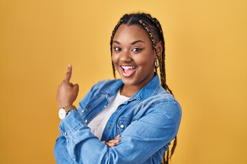 Wall Mural - African american woman with braids standing over yellow background with a big smile on face, pointing with hand finger to the side looking at the camera.