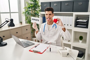 Canvas Print - Young hispanic doctor man supporting organs donations sticking tongue out happy with funny expression.