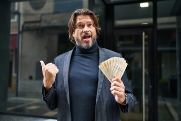 Canvas Print - Handsome middle age man holding 100 danish krone banknotes pointing thumb up to the side smiling happy with open mouth