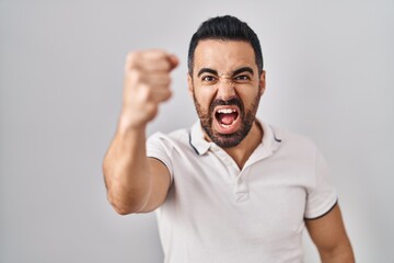 Sticker - Young hispanic man with beard wearing casual clothes over white background angry and mad raising fist frustrated and furious while shouting with anger. rage and aggressive concept.