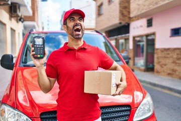 Wall Mural - Young hispanic man with beard wearing delivery uniform and cap holding dataphone angry and mad screaming frustrated and furious, shouting with anger looking up.