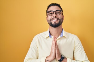 Canvas Print - Hispanic young man wearing business clothes and glasses praying with hands together asking for forgiveness smiling confident.