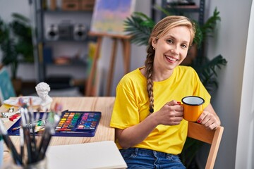 Sticker - Young blonde woman artist drinking coffee sitting on floor at art studio