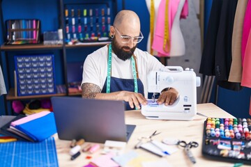 Poster - Young bald man tailor using sewing machine and laptop at clothing factory