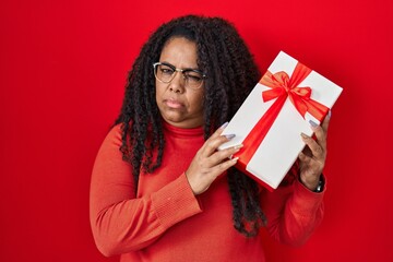 Sticker - Plus size hispanic woman holding gift skeptic and nervous, frowning upset because of problem. negative person.