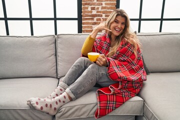 Canvas Print - Young woman sitting on the sofa drinking a coffee at home smiling doing phone gesture with hand and fingers like talking on the telephone. communicating concepts.