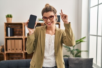 Canvas Print - Young woman working at consultation office holding smartphone surprised with an idea or question pointing finger with happy face, number one