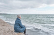 © IrinaK - Senior woman sitting on sea coast on cold cloudy day.