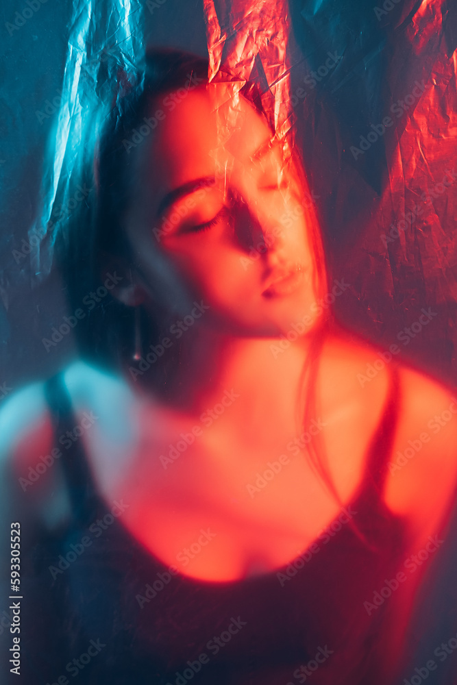 Light therapy. Facial aesthetic. Beauty skin treatment. Red color LED glow defocused relaxed woman face silhouette with closed eyes behind wrinkled transparent plastic film.