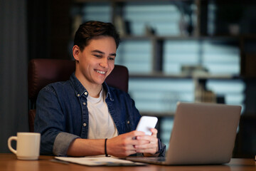 Aufkleber - Cheerful young man entrepreneur sitting at workdesk, using laptop, smartphone