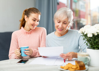 Happy senior mother and adult daughter doing financial paperwork while sitting at table in kitchen.