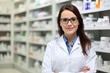 © aicandy - Elegant female pharmacist with a white lab coat and stethoscope standing confidently in front of medicine shelves with a friendly smile, generative ai