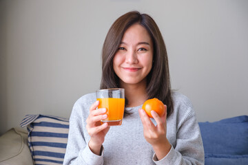 Wall Mural - Portrait image of a young woman holding an orange and a glass of fresh orange juice at home