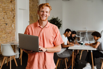 Wall Mural - Smiling man working on laptop computer while standing in office with his colleagues on a background